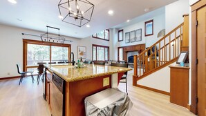 A spacious kitchen with granite countertops, a large island, and a dining area with chairs. There is a fireplace and a staircase leading to another level. - Pet-Friendly, Bright, Updated & On Golf Course! Private Game Room and Hot Tub (Mammoth Lakes)