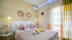 A neatly made bed with a beige bedspread, two wall-mounted mirrors, a bedside table with a vase of flowers, and a window with sheer curtains. - Villa Bella Luna (Zakynthos)