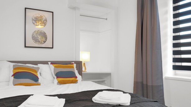 A hotel room with a bed, pillows, a framed artwork, a nightstand, a lamp, and a window with blinds.