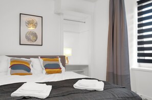 A hotel room with a bed, pillows, a framed artwork, a nightstand, a lamp, and a window with blinds. - Blackie's Neuk Ideal For Uni / West End (Dundee)
