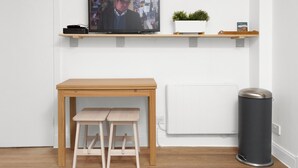 A room with a wooden table, two stools, a television on a shelf, a potted plant, and a trash can.