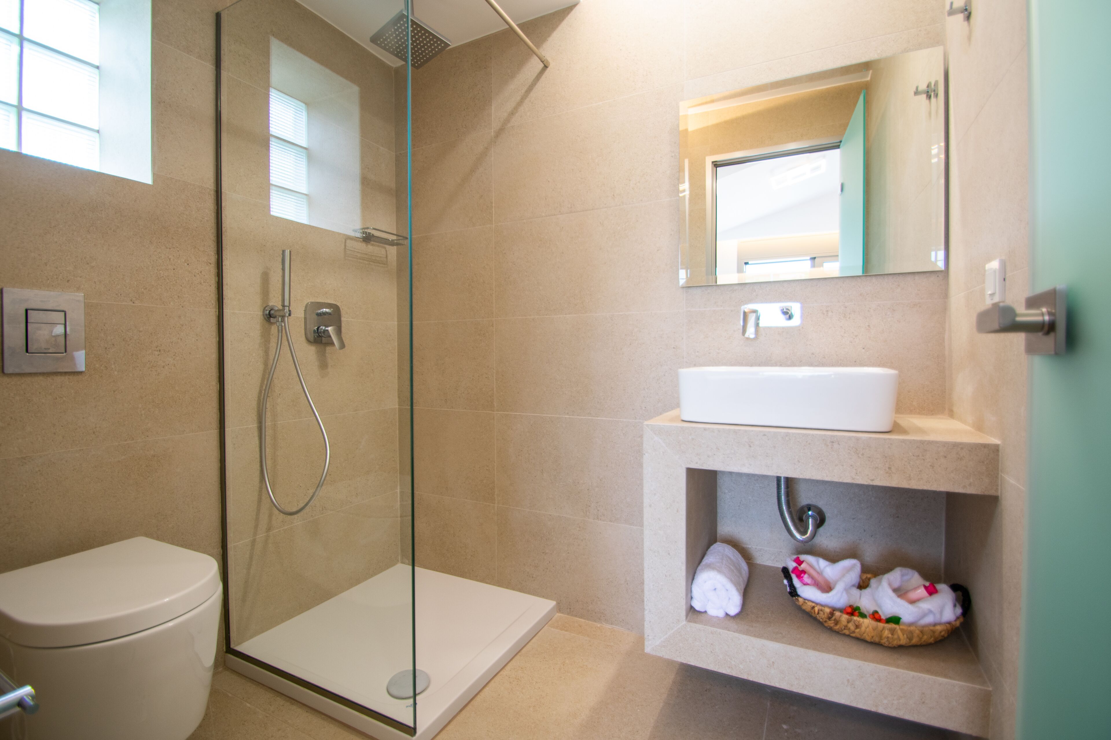 Ein modernes Badezimmer mit einer Duschkabine, einer weißen Toilette, einem Waschbecken mit Spiegel darüber und einem Ablagefach mit Handtüchern.