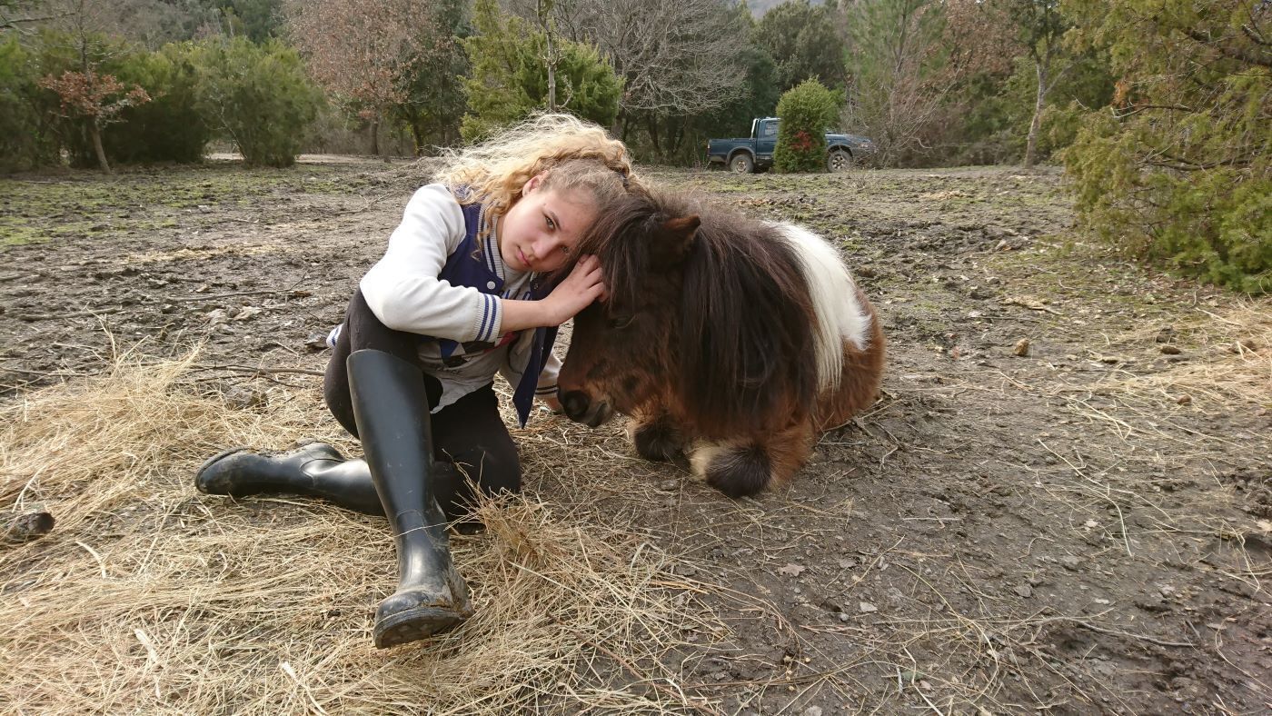 A person in rubber boots and a jacket hugging a pony with a long mane.