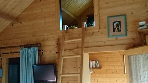 Interior de una cabaña de madera con habitación en entrepiso, un televisor montado en la pared y una pequeña mesa con una silla.