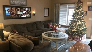 A cozy living room with a dark brown sectional sofa, a round coffee table with candles, a Christmas tree, and a 'Merry Christmas' banner. - Hometown Haven in Glens Falls, NY (between Lake George & Saratoga) (Glens Falls)