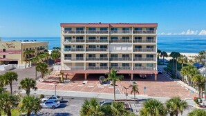 Un hôtel à plusieurs étages, doté d’un toit rouge et de balcons, situé près d’une plage bordée de palmiers et disposant d’un parking.