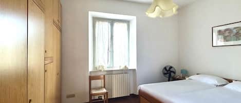 Un dormitorio con una cama, una silla, un ventanal, un radiador y un ventilador.