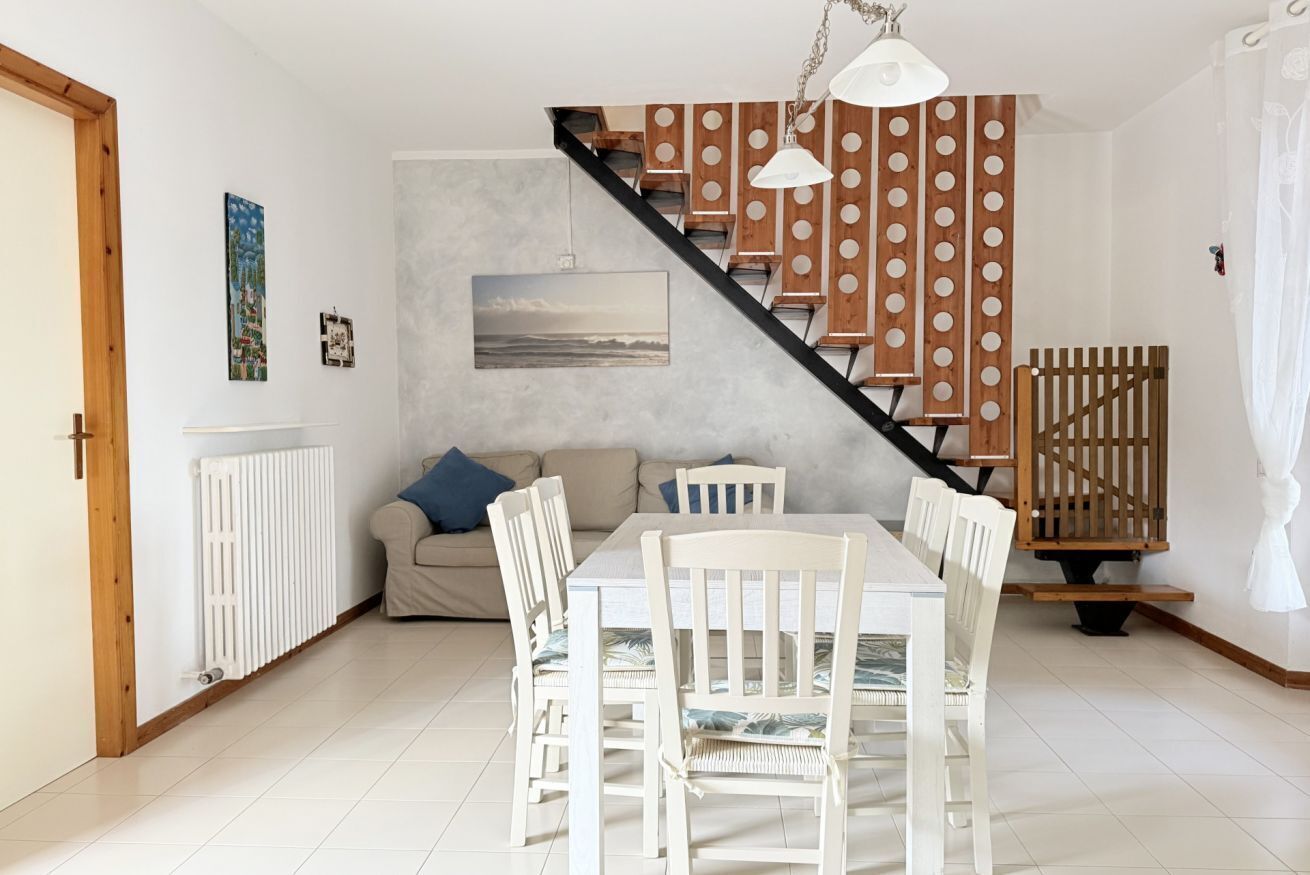 En matplats med ett vitt bord och stolar, en trappa med trästeg och ett svart räcke, ett vitt element och en beige soffa med blå kuddar.