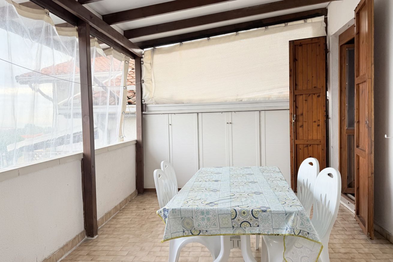 A covered patio with a table and chairs, a wooden door, and a window with a white blind.