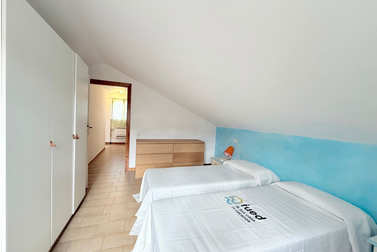 A bedroom with two beds, a wooden headboard, a nightstand with a lamp, and a view of a hallway.