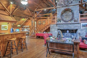 A rustic interior with a stone fireplace, wooden beams, a bar area with stools, and a staircase leading to an upper level.