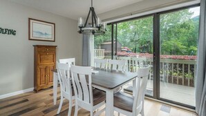A dining area with a table and chairs, a wooden cabinet, a chandelier, and a large sliding glass door leading to a balcony with a view of trees and a gazebo. - Fairway Villa 503 at Rumbling Bald (Lake Lure)