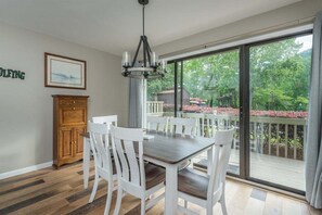 Uma área de refeições com mesa e cadeiras, um armário de madeira, um candelabro e uma grande porta de vidro deslizante que dá para uma varanda com vista para árvores e um gazebo.