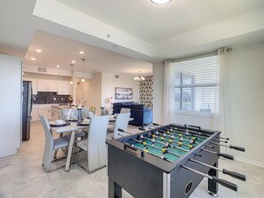 A modern living room with a foosball table, a dining area with a table and chairs, and a kitchen in the background.