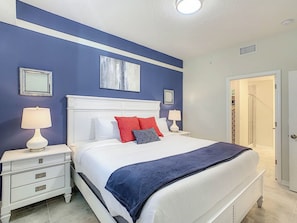 A bedroom with a white bed, red and blue pillows, a white nightstand, and a blue accent wall.