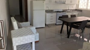Een moderne keuken met witte kasten, een koelkast en een fornuis. Er is een eettafel met stoelen en een bank met een decoratief hoesje.