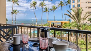 A balcony with a table set for breakfast overlooking a beach with palm trees and boats.