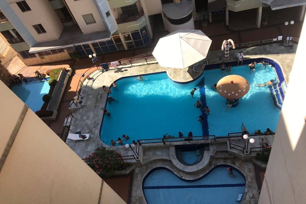 An indoor swimming pool with multiple lanes, a central water slide, and a surrounding deck area with seating and umbrellas.