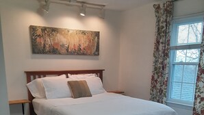 Ein Schlafzimmer mit Bett, Nachttisch, einem Gemälde an der Wand und einem Fenster mit Vorhängen.