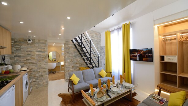 A modern living room with a grey sofa, wooden dining table set for a meal, a staircase, and a kitchen area with a washing machine. - B4DD Opportunity (París)