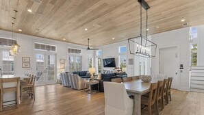 Un vaste espace de vie doté d’un plafond en bois, de grandes fenêtres et d’un coin repas avec une longue table et des chaises.