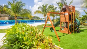 A playground with a swing set and slide, surrounded by palm trees and a pool.