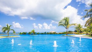 A large outdoor swimming pool with multiple fountains, surrounded by palm trees and a clear sky.