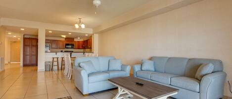 A spacious living room with a light blue sectional sofa, a wooden coffee table, and a kitchen area in the background.