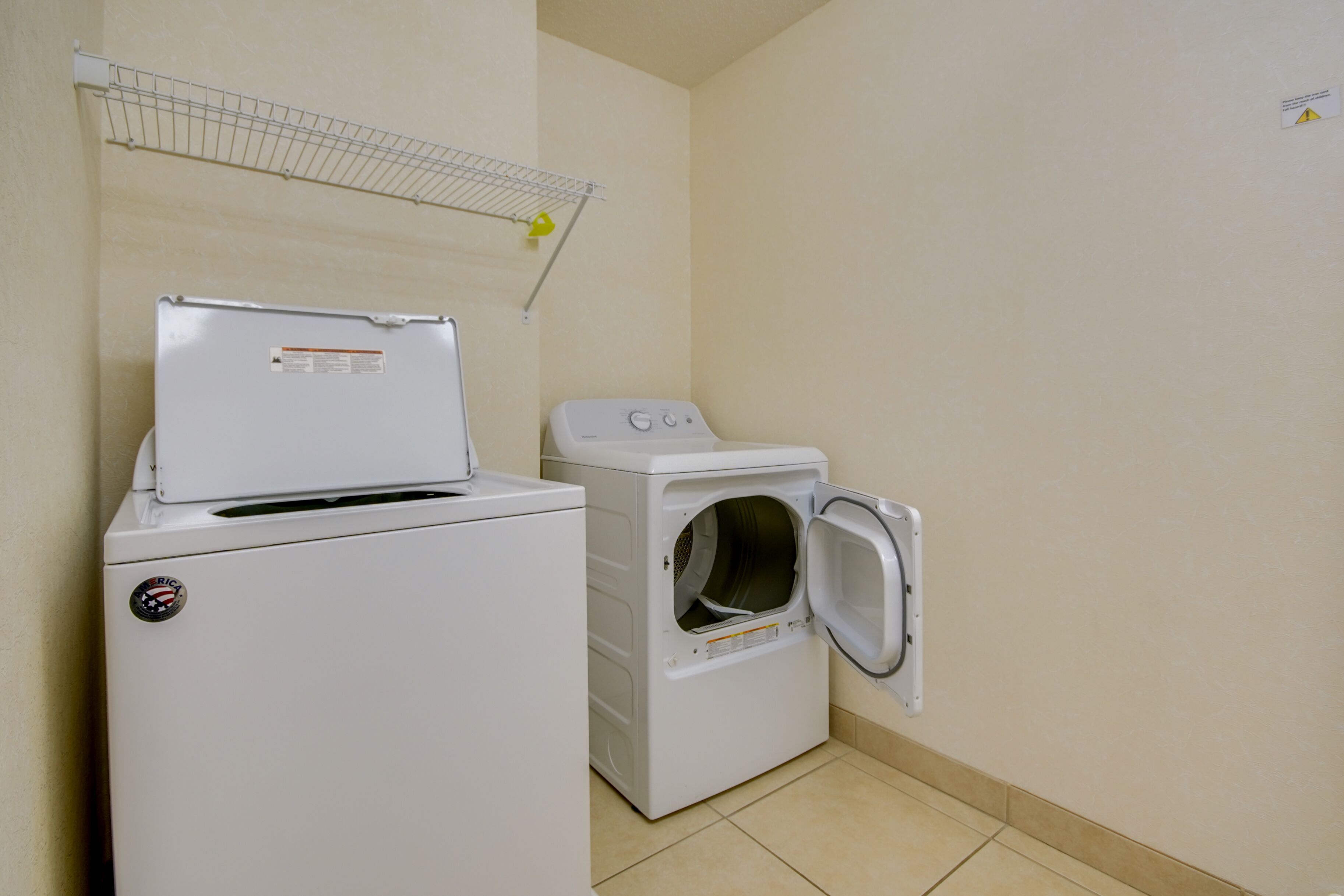 Un espace buanderie comprenant un lave-linge et un sèche-linge, une étagère en métal au-dessus du sèche-linge et un sol carrelé.