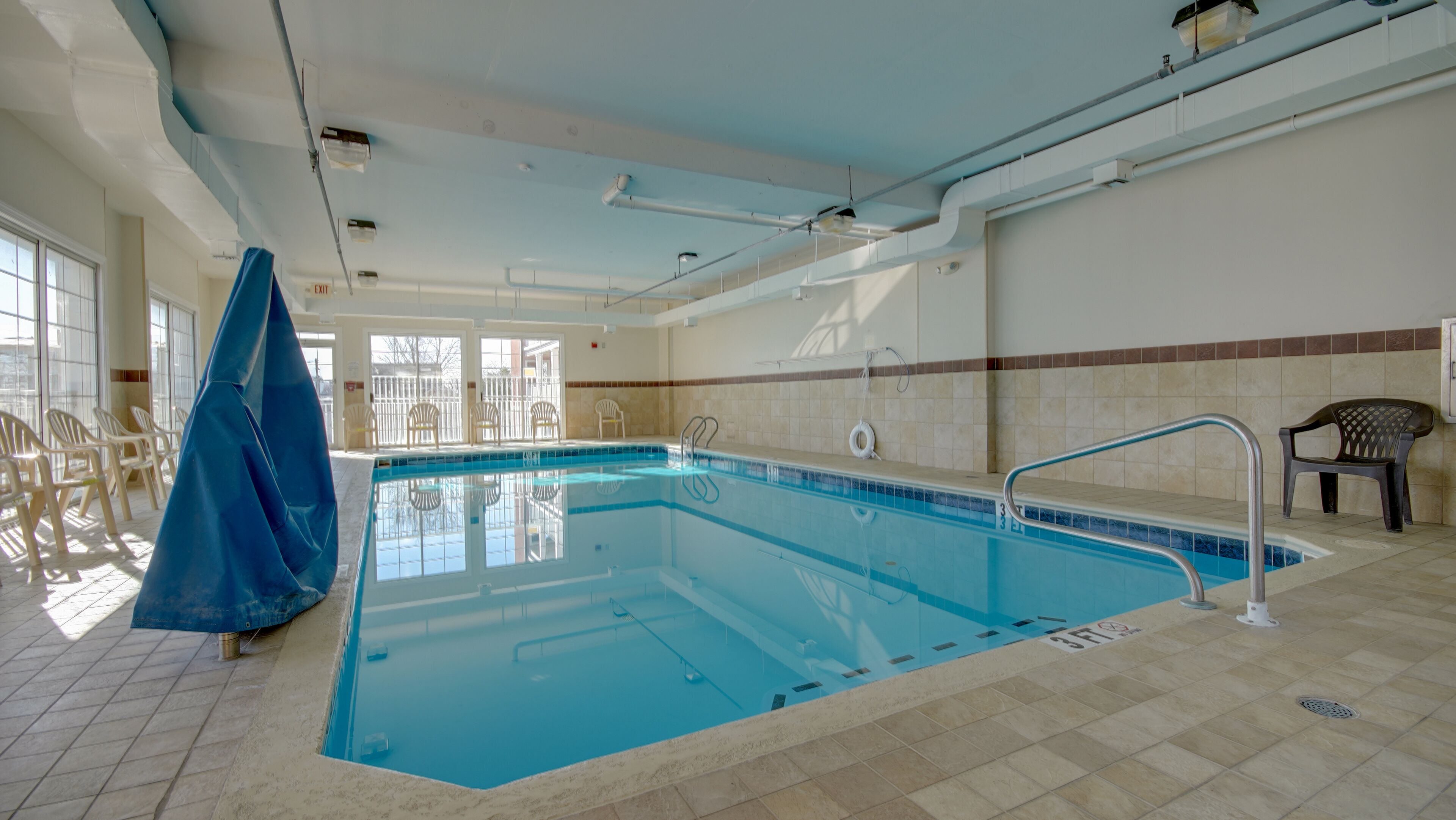 An indoor swimming pool with clear water, tiled walls, and a tiled floor. There are chairs and a covered area to the left.
