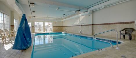 An indoor swimming pool with clear water, tiled walls, and a tiled floor. There are chairs and a covered area to the left.