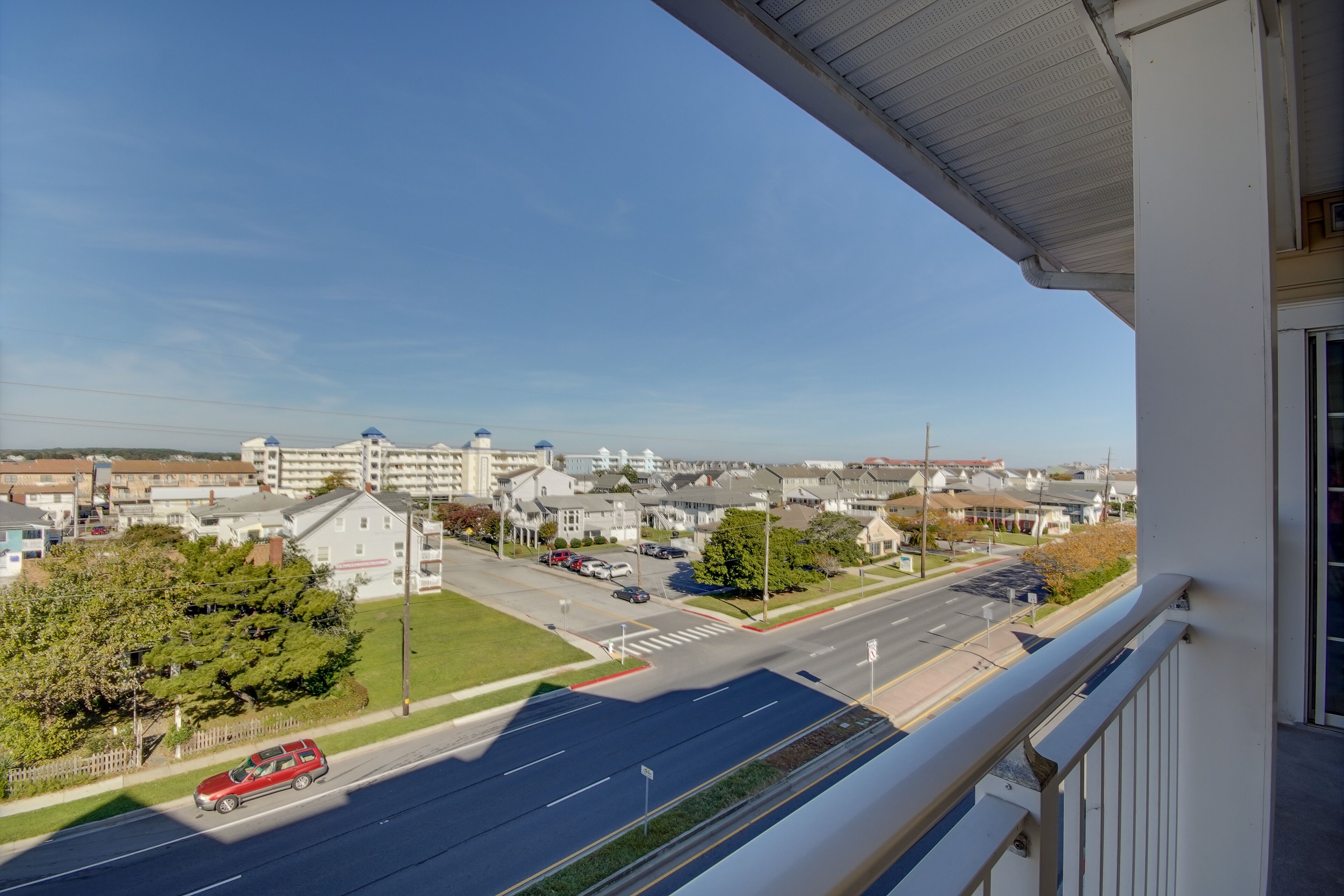 A view from a balcony overlooking a street with buildings, a road, and a red car.