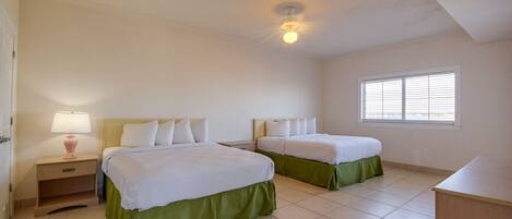 A clean, well-lit bedroom with two beds, a nightstand with a lamp, a window with blinds, and a ceiling fan.