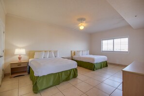 A clean, well-lit bedroom with two beds, a nightstand with a lamp, a window with blinds, and a ceiling fan. - Americana Condos 10th street (Ocean City)