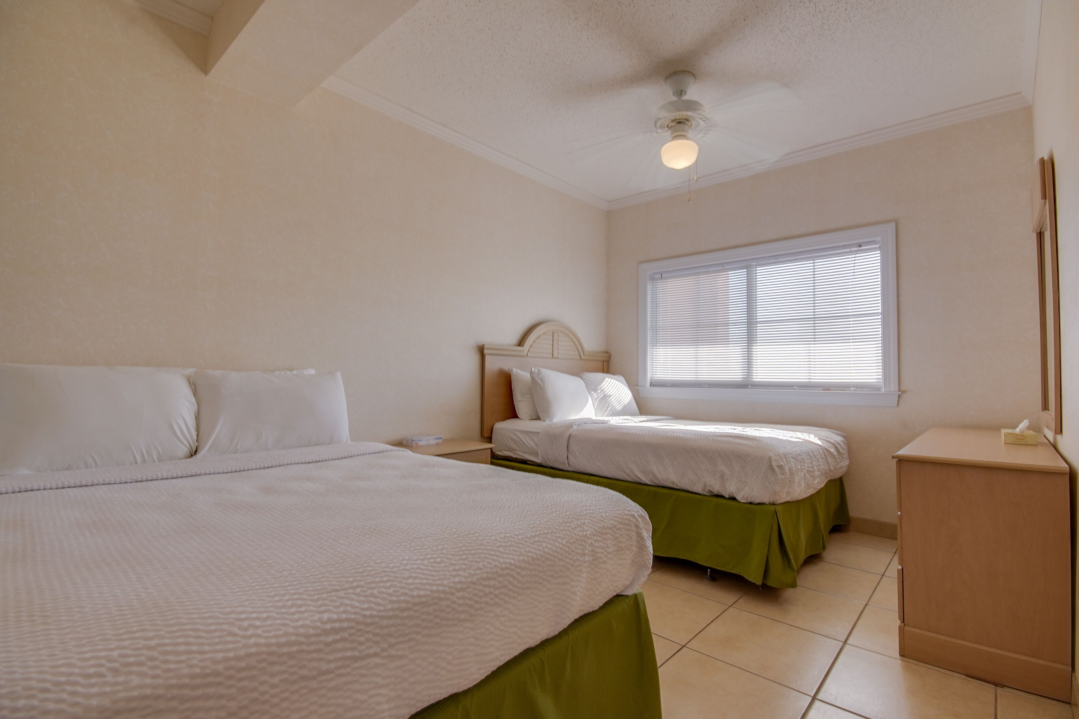 A bedroom with two beds, a window with blinds, a ceiling fan, and a small wooden cabinet.