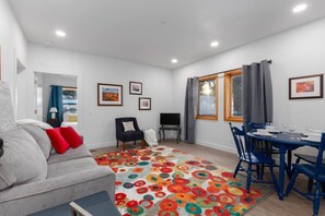 Ein Wohnzimmer mit grauem Sofa, roten Kissen, einem farbenfrohen Teppich, einem schwarzen Sessel, einem Esstisch mit blauen Stühlen und gerahmten Bildern an den Wänden.