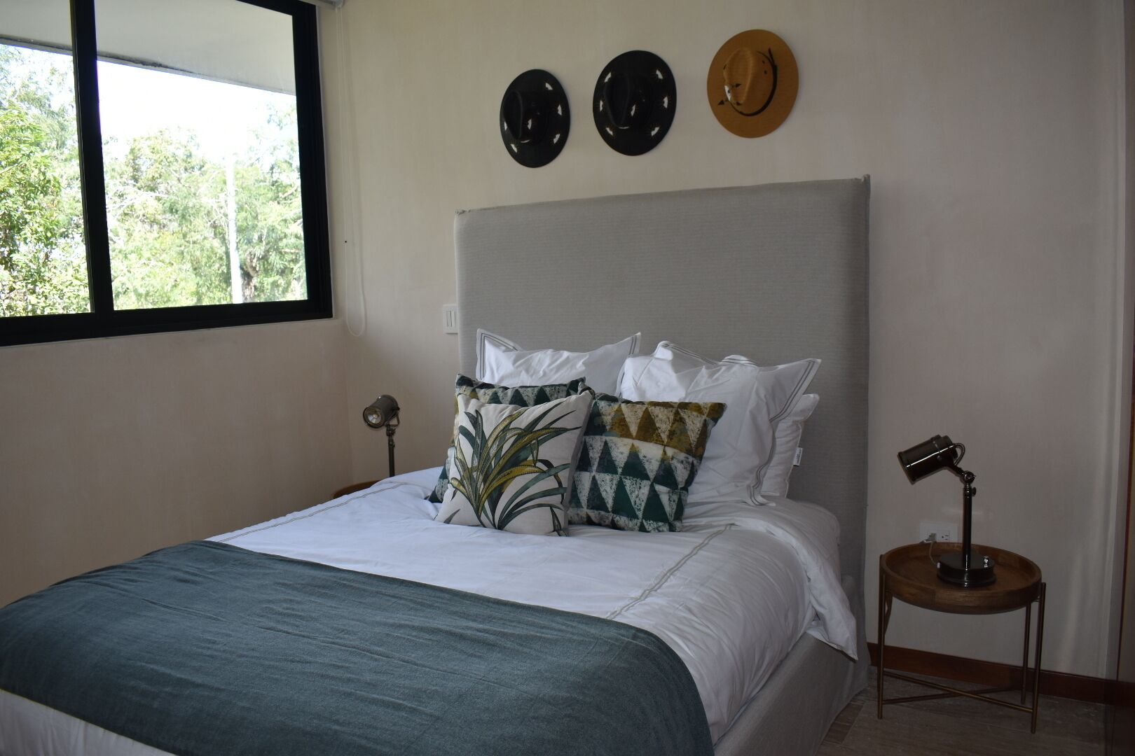 Una cama bien hecha con un edredón azul, almohadas blancas y una almohada estampada. Encima de la cama, hay tres sombreros colgados en la pared. Una mesita de noche con una lámpara está al lado de la cama.