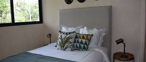 Una cama bien hecha con un edredón azul, almohadas blancas y una almohada estampada. Encima de la cama, hay tres sombreros colgados en la pared. Una mesita de noche con una lámpara está al lado de la cama.