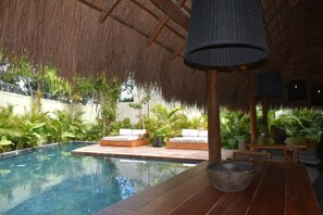 Ein Poolbereich mit Holzterrasse, einem Reetdach und einer Liege.