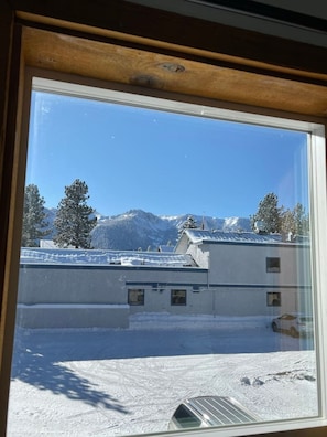 Blick aus dem Fenster auf ein Gebäude, mit verschneitem Boden, Bäumen und Bergen im Hintergrund.