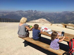 Een gezin van drie personen aan een picknicktafel, met een schilderachtig uitzicht op de bergen op de achtergrond.