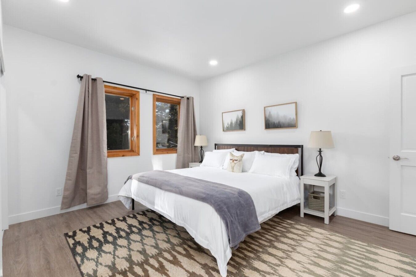 A bedroom with a bed, two bedside tables, a window with curtains, a rug, and two wall-mounted pictures.