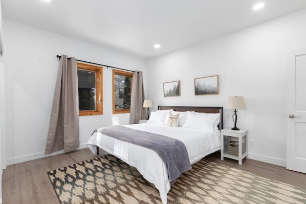 A bedroom with a bed, two bedside tables, a window with curtains, a rug, and two wall-mounted pictures.