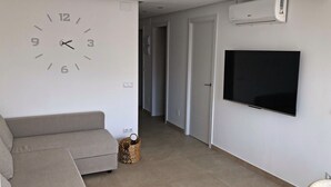 A living room with a beige sofa, a wall clock, a flat-screen TV, and an air conditioning unit. - Cristian's Apartment 2 (Benidorm)