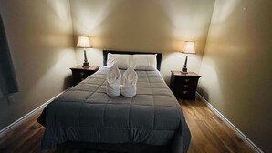 Una cama bien hecha con una colcha gris oscuro, dos toallas blancas y dos pantuflas blancas colocadas sobre ella. Hay dos mesitas de noche con lámparas, una a cada lado de la cama.