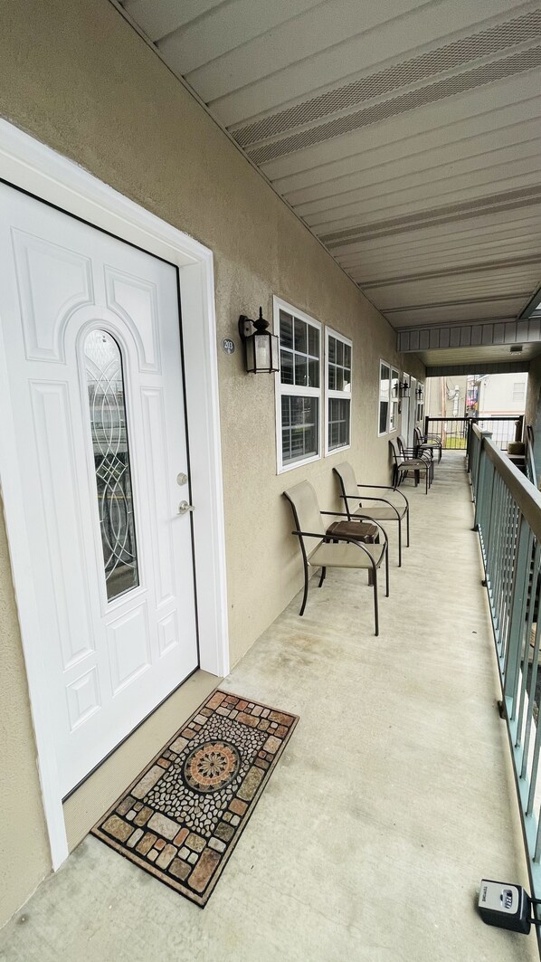 A covered porch with a white door, a tiled doormat, and a row of chairs.