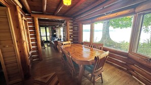 A log cabin interior with a large wooden dining table, chairs, and a view of a lake through large windows.