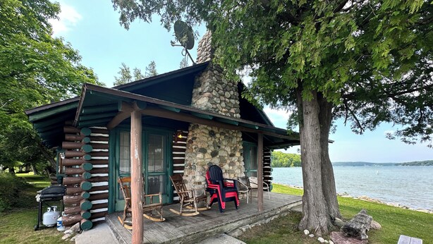 A rustic cabin with a stone wall and wooden logs, a porch with rocking chairs, and a view of a lake.