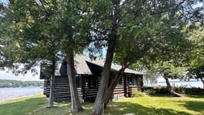 A log cabin with a sloped roof, surrounded by trees and a grassy area.