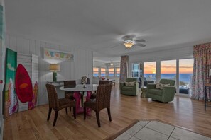 Uma área de refeições com mesa redonda e cadeiras, ventilador de teto e vista para o oceano ao pôr do sol.